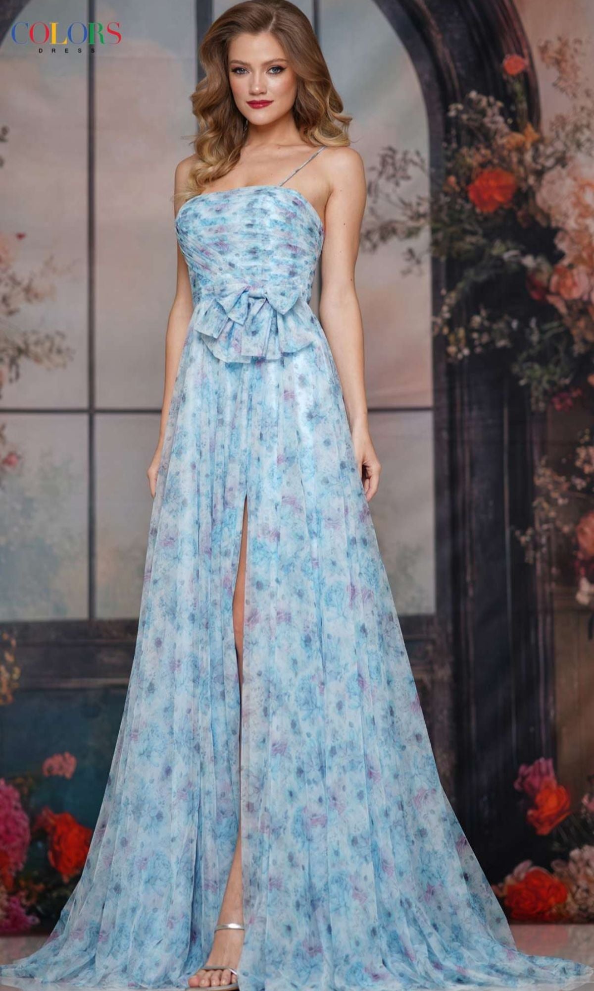 Woman in a strappy long blue print prom dress, Colors Dress 3812, featuring a front slit and thin straps. She stands indoors by a window with a floral backdrop, showcasing the elegance of this standout style.